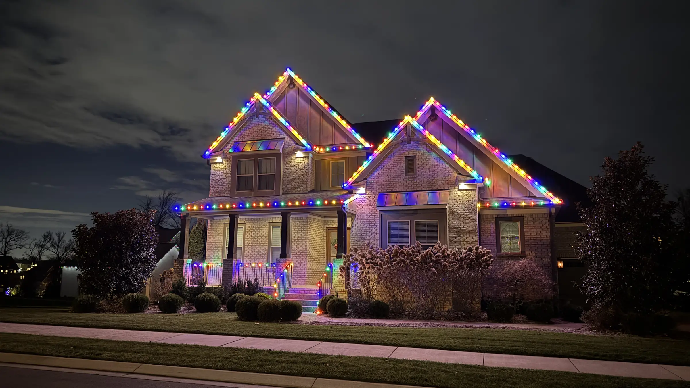 A two story home with red, green and warm white Christmas lights along the rooflines and porch