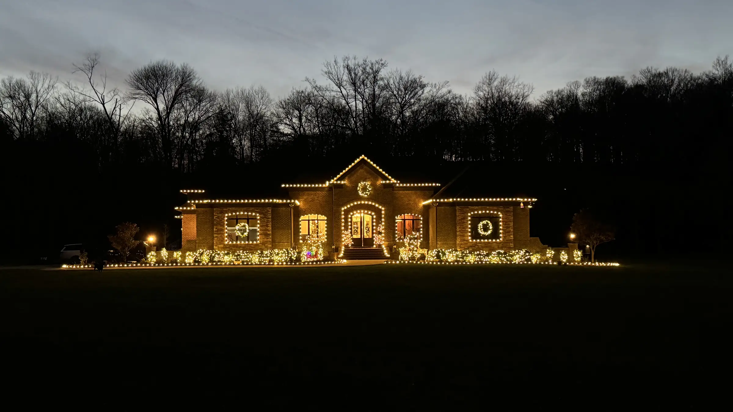 A single story home outlined in warm white along the full roofline, with lit wreaths in every window and lit planting beds