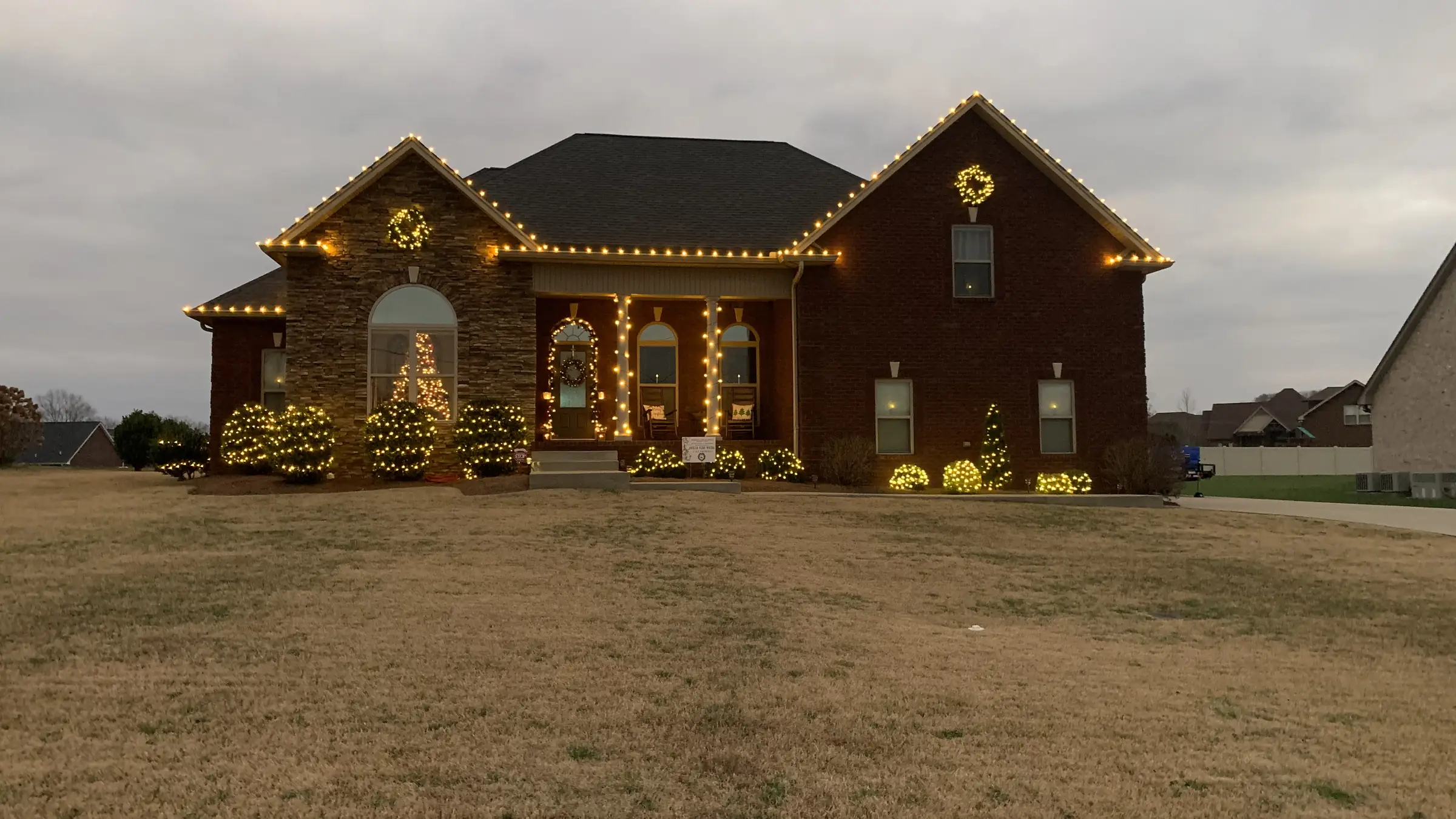 A brick home in Greenbrier, Tennessee with warm white bulbs running the full roofline, lit wreaths and lit trees at the porch