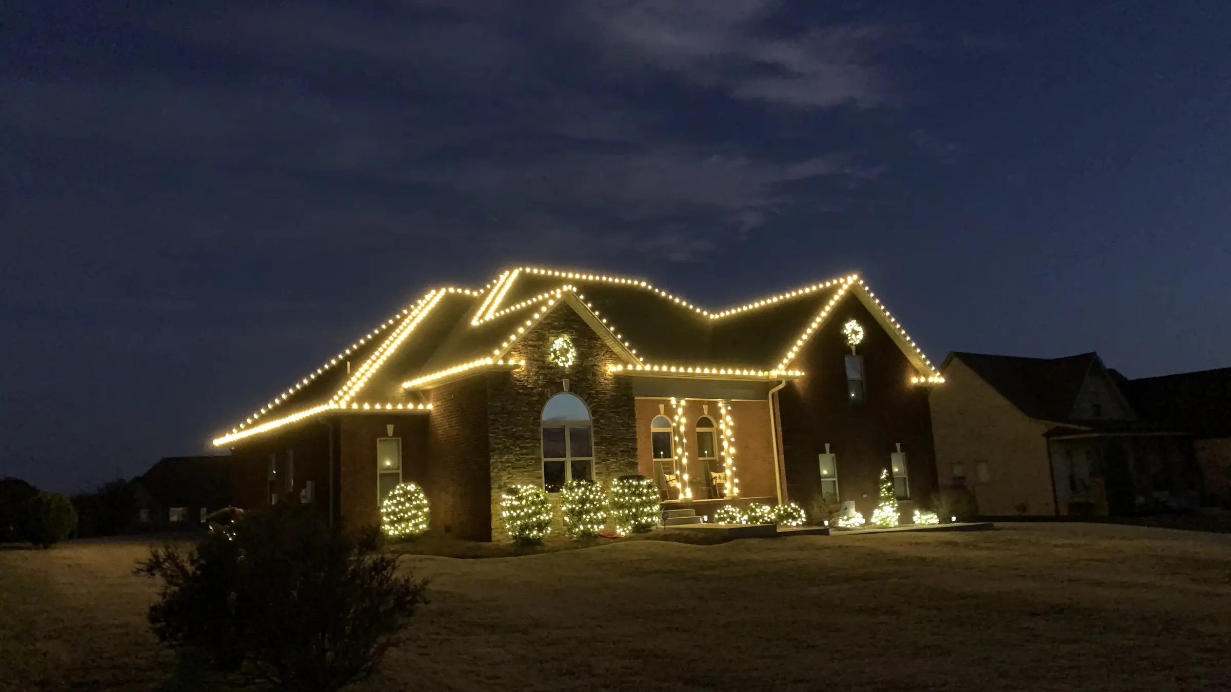 A brick home in Greenbrier, Tennessee outlined in warm white along every roofline and gable, with lit wreaths and lit shrubs at dusk