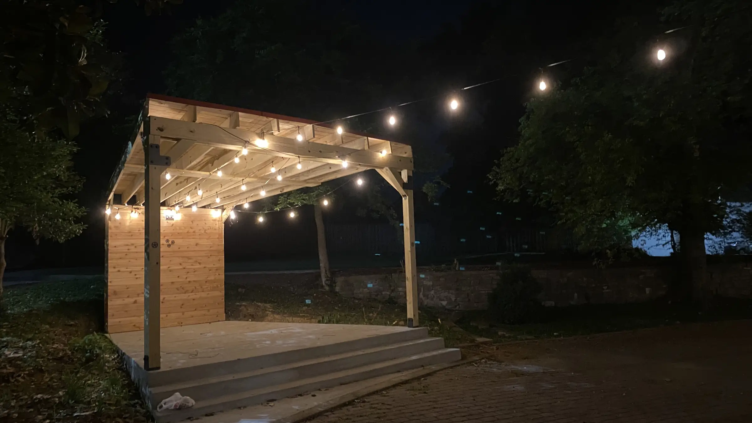 A wooden outdoor stage in Gallatin, Tennessee with string lights along its roof beams and a strand running overhead