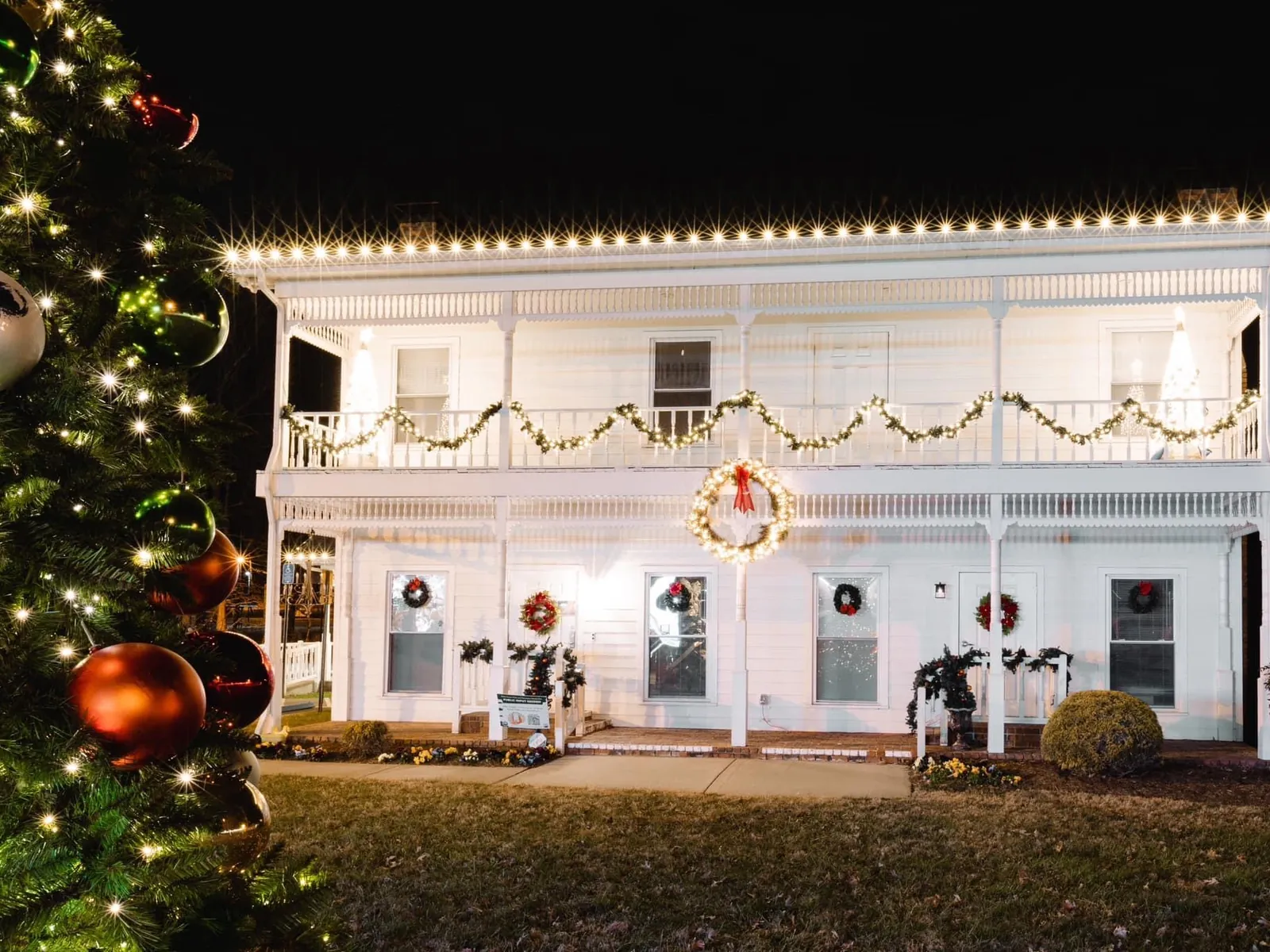 A white two story house with garland and wreaths across both porch levels and warm white lights along the eaves