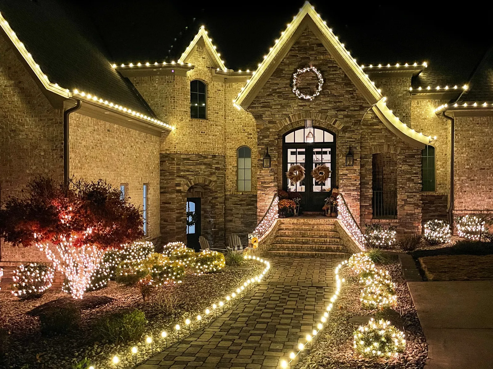 A stone house with warm white lights along the rooflines and a lit path curving to the front door