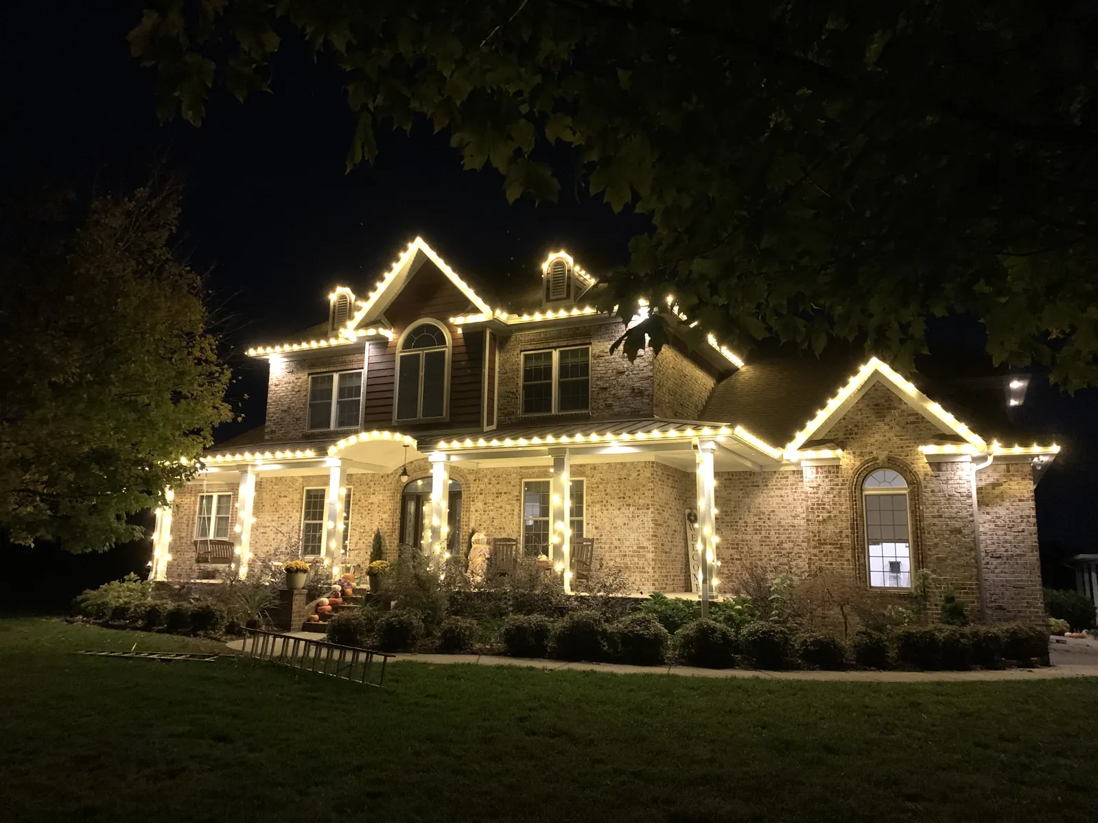 A brick two story home under mature trees with warm white lights along the rooflines and dormers