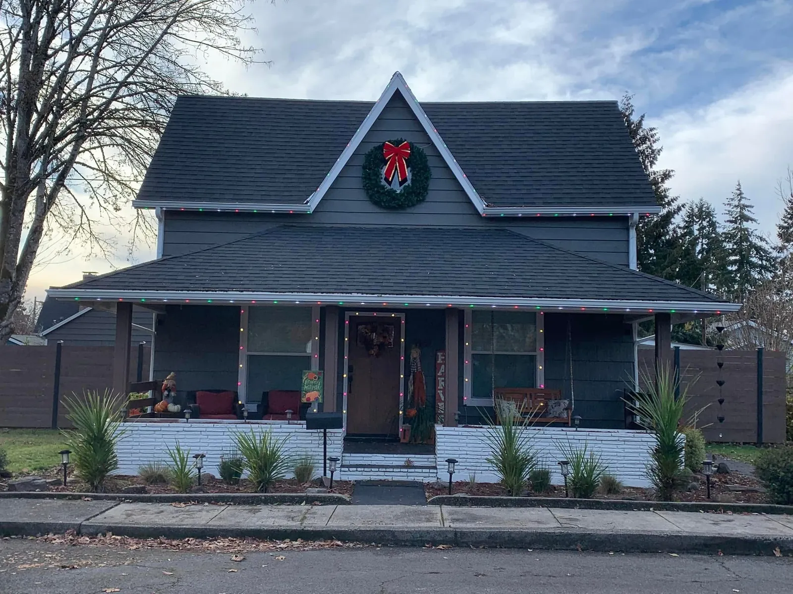 A gray clapboard house with a red bow wreath at the gable peak and lights along the porch roof